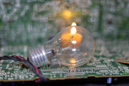 An incandescent lamp against the background of a burning candle and electronic circuit boards. Blackout due to the war in Ukraine