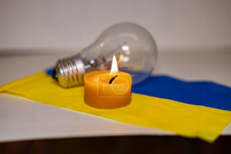 Burning candle on the background of a light bulb and the Ukrainian flag. Blackout due to the war in Ukraine