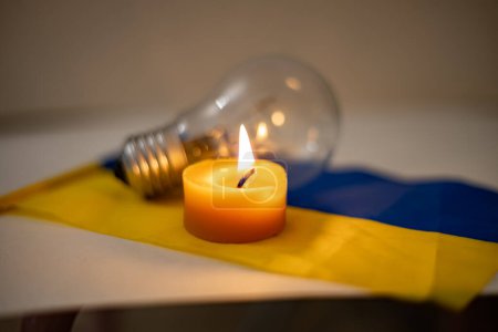 Burning candle on the background of a light bulb and the Ukrainian flag. Blackout due to the war in Ukraine