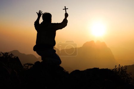 Téléchargez les photos : Silhouette de Jeune homme priant et tenant la croix chrétienne pour adorer Dieu au coucher du soleil. Religion chrétienne concept arrière-plan. - en image libre de droit