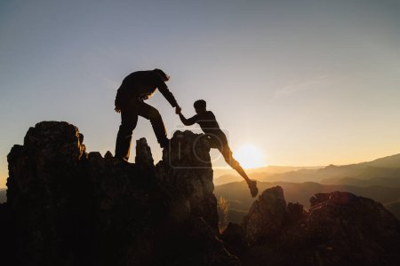 Foto de Silhouette of Teamwork of two men hiker helping each other on top of mountain climbing team. Teamwork friendship hiking help each other trust assistance silhouette in mountains, sunrise. - Imagen libre de derechos