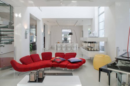Modern interior of luxury private house. Spacious living room with red sofa, piano, white table. Metal and glass spiral staircase.