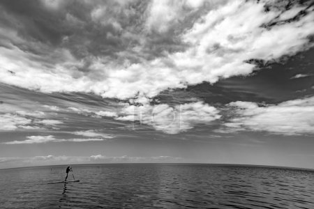 Téléchargez les photos : Paysage marin pittoresque de mer calme et ciel nuageux. Un homme flottant sur la planche à souper. Noir et blanc. - en image libre de droit