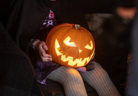 Téléchargez les photos : Lanterne de citrouille Halloween avec visage effrayant sur les genoux de l'enfant. - en image libre de droit