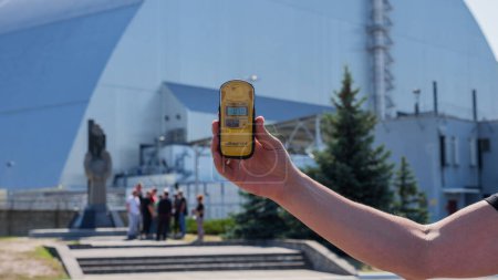 Foto de Zona de exclusión. Ucrania. Pripyat. 26 de agosto de 2019. Dosimetro en mano sobre fondo cuarto reactor central nuclear de Chernobyl bajo nuevo sarcófago seguro. Monumento a los liquidadores accidente de Chernobyl - Imagen libre de derechos