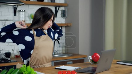 Téléchargez les photos : Femme au foyer fatiguée dans le tablier à la maison cuisine étire les muscles tendus après longtemps étudier à distance webcam en ligne conférence téléphonique webinaire cours ordinateur portable, mauvaise posture nécessitant un traitement - en image libre de droit