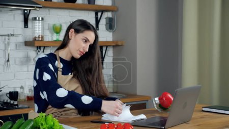 Téléchargez les photos : Femme femme au foyer en tablier beige dit professeur chef recherche recette culinaire dans étude ordinateur portable en ligne téléconférence webcam ordinateur portable, cours de webinaire d'enseignement à distance dans la cuisine à la maison - en image libre de droit