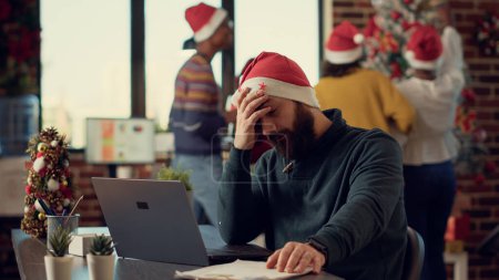 Photo for Stressed person trying to work in festive decorated office, being interrupted by noisy people celebrating christmas season. Festive colleagues making noise, disturbing man with santa hat. Tripod shot. - Royalty Free Image