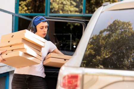 Téléchargez les photos : Un coursier transportant des pizzas au bureau en voiture, une jeune femme tenant des boîtes empilées. Employé du service de livraison de pizzeria dans les écouteurs transportant le déjeuner, personne sortant la restauration rapide du véhicule - en image libre de droit