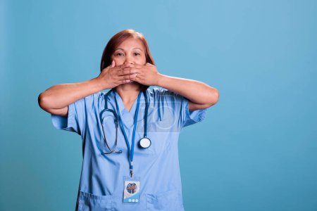 Photo for Elderly physician nurse in blue uniform covering mouth with hands, speak no evil, three wise monkeys concept. Medical assistant working at health care expertise planning disease treatment - Royalty Free Image