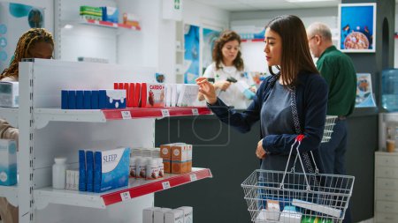 Foto de Cliente asiático mirando cajas de medicamentos y pastillas botellas en los estantes, comprobar los productos farmacéuticos para comprar productos médicos y suplementos. Mujer que lee el prospecto de medicamentos. - Imagen libre de derechos