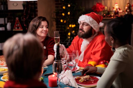 Photo for Man in santa claus costume proposing christmas toast with sparkling wine glass, celebrating holiday with family at home feast. Xmas celebration, people gathering at dinner table - Royalty Free Image