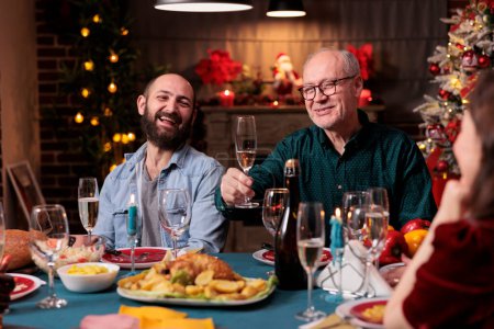 Photo for Elderly man proposing christmas toast at family gathering, laughing, holding glass with sparkling wine at festive dinner. People celebrating xmas holiday, drinking at new year home party - Royalty Free Image