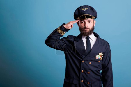 Photo for Serious airplane pilot saluting, wearing uniform and hat front view portrait, plane captain looking at camera. Aviation academy aviator with airline wings badge on jacket - Royalty Free Image