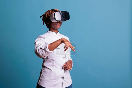 Photo for Professional female chef using virtual reality goggles in studio shot. Afro american cook wearing uniform excitedly in a simulation of culinary creations against blue background. - Royalty Free Image