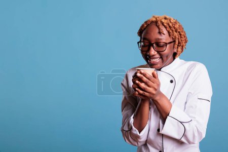 Téléchargez les photos : Cuisinière détendue profitant d'une tasse de café préféré pendant l'heure du déjeuner. Chef professionnel afro-américain portant un uniforme de cuisine sur fond bleu en studio. - en image libre de droit