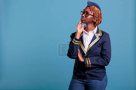 Photo for Female african american flight attendant thinking, wondering. Laughing hair, young stewardess, wearing glasses medium shot, posing with thoughtful expression for studio portrait. - Royalty Free Image