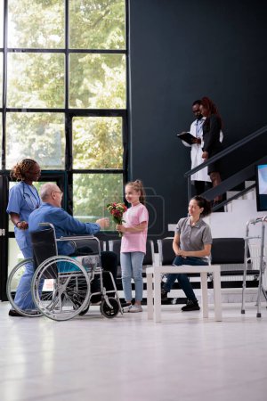Photo for Little girl giving bouquet of flowers to disabled grandfather during checkup visit appointment in hospital waiting room. Patient in wheelchair discussing health care treatment with medical assistant - Royalty Free Image