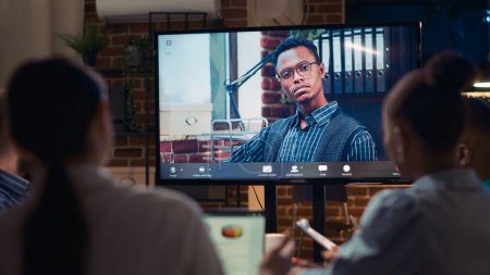 Líder de equipo remoto afroamericano hablando en videocall, pantalla de cerca, diversas personas hablando en videoconferencia por la noche. Trabajadores de oficina charlando en teleconferencia en espacio de coworking