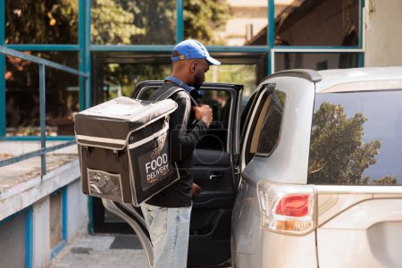 Téléchargez les photos : Jeune homme livrant commande de nourriture de bureau en voiture. Emporter déjeuner livreur tenant sac à dos thermique avec repas, debout près du véhicule à l'extérieur, vue de côté plan moyen - en image libre de droit
