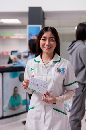 Foto de Sonriente farmacéutico asiático que sostiene la caja de vitaminas en la farmacia medicinal, mostrando el paquete de medicamentos y suplementos. Pastillas publicitarias y medicamentos de los estantes de farmacia. - Imagen libre de derechos