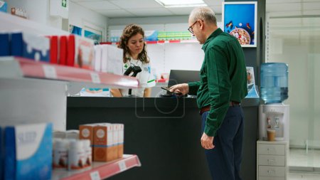 Photo for Elderly customer buying pills for prescription medicine at drugstore counter paying with mobile phone nfc payment. Senior man talking to pharmacist about medicaments and vitamins. - Royalty Free Image