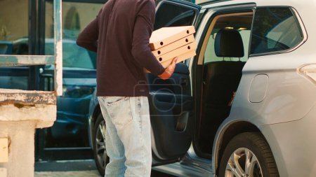 Téléchargez les photos : Courrier homme afro-américain laissant la voiture pour livrer des boîtes de pizza à la porte d'entrée, en utilisant l'automobile donnant commande de repas pizzeria au client. Livreur avec sac à dos sonnant cloche pour livrer de la nourriture. - en image libre de droit