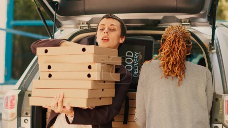 Téléchargez les photos : Livreur afro-américain prenant des colis dans le coffre de la voiture, donnant des piles de boîtes à pizza à la cliente. Courrier de nourriture rapide livrant grande commande de pizzeria à emporter à la femme. Coup de main. - en image libre de droit