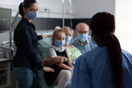 Téléchargez les photos : Infirmière afro-américaine répondant aux questions de la famille des femmes récupérées à l'hôpital du coronavirus. Joyeuse famille à l'écoute de bonnes nouvelles sur la santé de la grand-mère à la clinique de gériatrie. - en image libre de droit