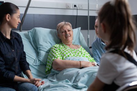 Photo for Smiling elderly woman under medical observation in hospital, chatting happily with girl. Relatives visiting senior lady during hospital stay. Happy older female patient with several people at - Royalty Free Image