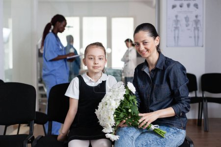 Photo for Young woman, little girl looking at camera, seated on medical clinic reception couches. Mother, daughter carrying flowers to hospitalized relative. Family members waiting in medical tower lobby. - Royalty Free Image