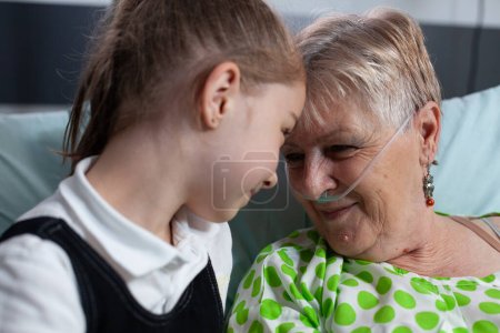 Family members smiling happily at sanatorium nursing home. Girl showing affection to elderly patient with breathing aids in hospital room. Grandmother grateful for granddaughter visit at medical