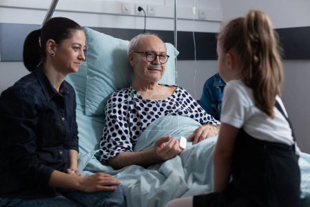 Photo for Elderly man lying in hospital bed chatting with visiting family members. Little girl talking to grandfather in elderly people medical clinic. Family living with old man resting in sanatorium bed. - Royalty Free Image