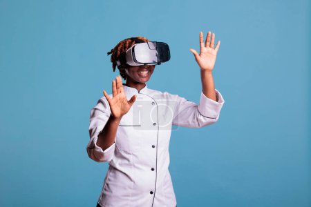 Photo for Funny female cook with VR goggles enjoys virtual reality experience on blue background in studio photo. Chef wearing uniform moving his hands while watching a simulation. - Royalty Free Image