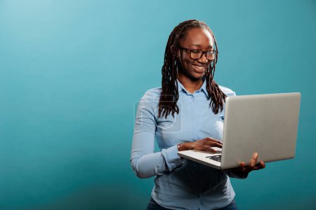 Foto de Cheerful happy woman having laptop and smiling heartily while browsing webpages on internet. Joyful pretty lady with handheld modern computer standing on blue background while surfing browser. - Imagen libre de derechos