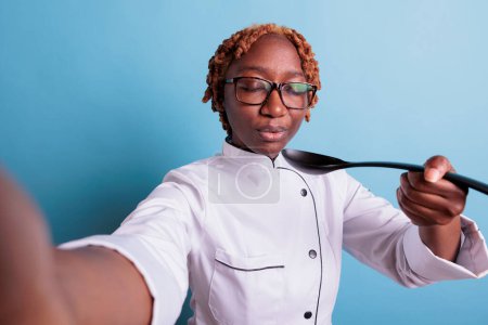 Téléchargez les photos : Femme chef afro-américaine soufflant cuillère avec soupe chaude. Cuisinière professionnelle les yeux fermés portant l'uniforme de cuisine en studio tourné isolé en bleu, concept de cuisine. - en image libre de droit