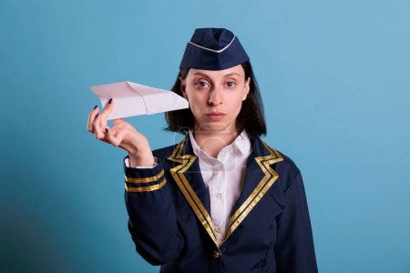 Photo for Stewardess holding paper plane, looking at camera, aviation academy flight attendant playing with airplane model. Air hostess looking at commercial jet toy, front view portrait - Royalty Free Image