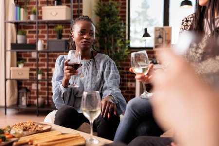 Foto de Mujer afroamericana bebiendo vino, escuchando, escuchando atentamente la conversación de amigos, hablando, discutiendo, charlando en casa, casa, fiesta del apartamento, reunión, reunión. - Imagen libre de derechos