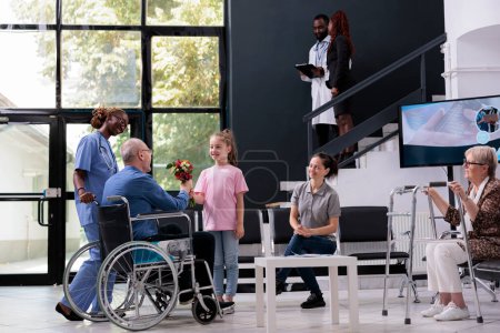 Photo for Granddaughter handing bouquet of flowers to grandfather in wheelchair after finishing medical examination in hospital waiting room. Elderly man discussing health care treatment with nurse - Royalty Free Image