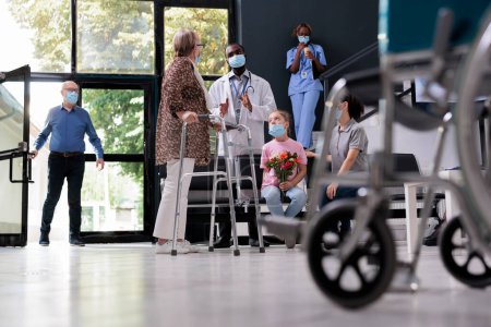 Photo for Elderly patient with walking frame discussing health care treatment with medic in hopsital waiting area, granddaughter holding flowers for grandmother. People with face mask against covid19 - Royalty Free Image