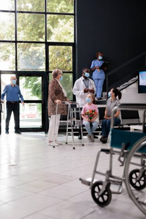 Photo for Senior woman with walking frame discussing disease diagnosis with doctor during checkup visit in hospital reception. People wearing medical protective face mask to prevent infection with coronavirus - Royalty Free Image