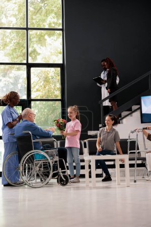 Photo for Granddaughter giving bouquet of flowers to disabled grandfather during checkup visit appointment in hospital waiting area. Patient in wheelchair discussing medication treatment with medical nurse - Royalty Free Image