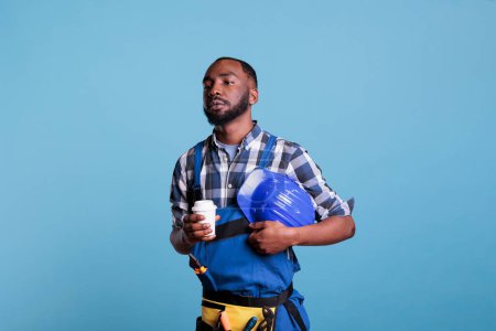 Photo for Tired construction worker drinking a cup of coffee at the end of his workday. Handyman in work uniform holding hard hat under his arm while enjoying a relaxing hot drink. - Royalty Free Image