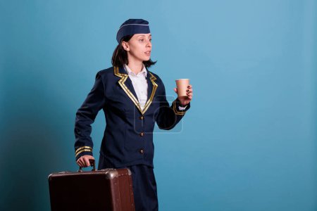 Photo for Flight attendant in professional aviation uniform running late, carrying suitcase. Stewardess with baggage walking in airport, woman holding cup of coffee, side view medium shot - Royalty Free Image