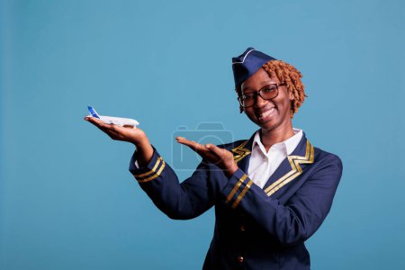 Téléchargez les photos : Une hôtesse afro-américaine souriante pointant du doigt un avion commercial tenant en main la publicité des compagnies aériennes. Un agent de bord portant l'uniforme d'un membre d'équipage en studio. - en image libre de droit