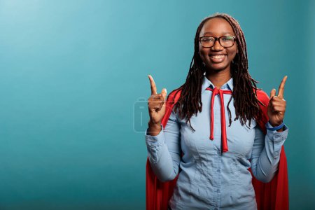 Photo for Confident and strong superhero woman wearing red cloak poiting fingers up while standing on blue background. Selfless brave justice defender posing for camera while gesturing fingers up. Studio shot - Royalty Free Image