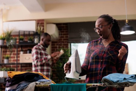 Foto de Mujer afroamericana usando plancha de vapor en casa, impresionada al descubrir cómo funciona. Ama de casa limpiando y haciendo todas las tareas mientras el hombre está leyendo un libro en segundo plano, ropa de planchar para adultos femeninos - Imagen libre de derechos
