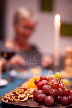 Photo for Nuts and grapes sitting on wooden plate during elderly couple festive dinner. Senior couple sitting at the table in kitchen, talking, enjoying the meal, celebrating their anniversary in the dining - Royalty Free Image