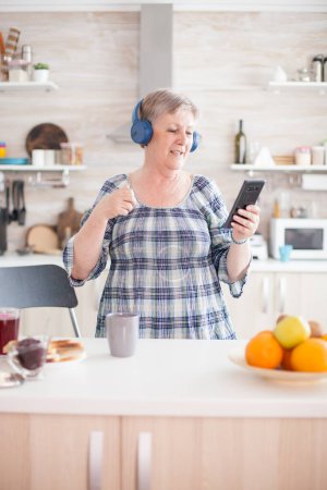 Foto de Mujer mayor relajada escuchando música en auriculares durante el desayuno en la cocina. Personas mayores bailando, estilo de vida divertido con tecnología moderna - Imagen libre de derechos