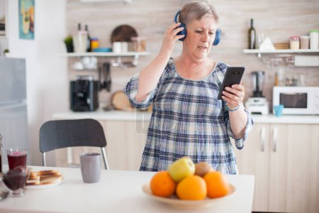 Foto de Mujer mayor relajada escuchando música en los auriculares durante el desayuno en la cocina. Personas mayores bailando, estilo de vida divertido con tecnología moderna - Imagen libre de derechos